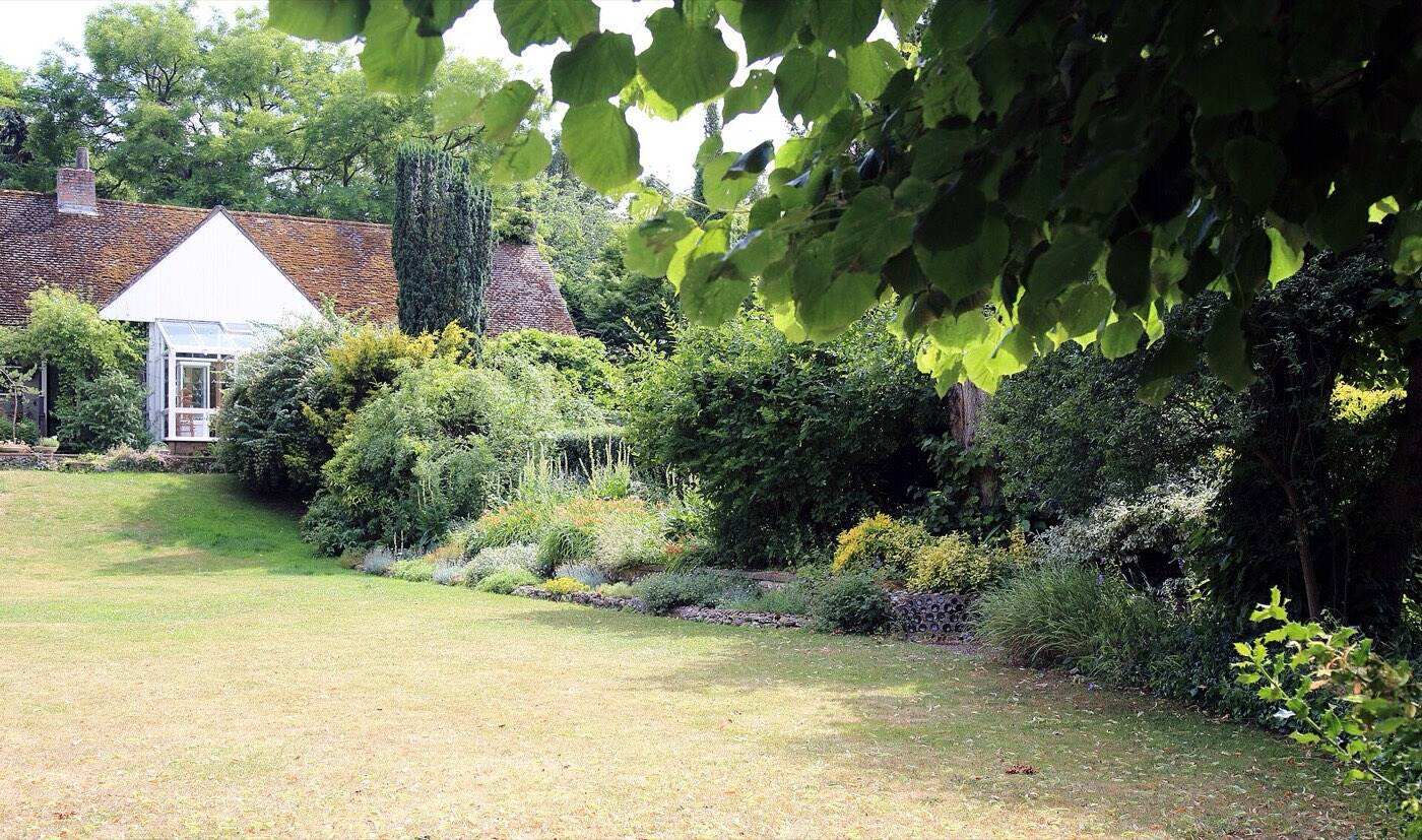 Garten mit Rasenfläche, dicht bepflanzten Staudenbeeten und Gehölzen vor einem Wohnhaus